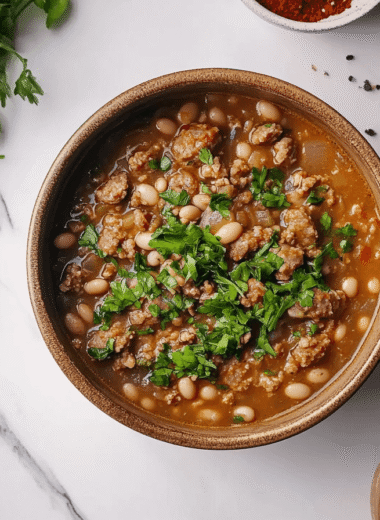A hearty bowl of white bean and sausage soup garnished with fresh herbs, served in a rustic ceramic bowl. The bowl sits on a pristine white marble kitchen island, captured from an overhead perspective in natural light, emphasizing its rich textures.